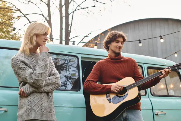 Two young men share a moment of joy in a charming outdoor space during fall. One plays the guitar while the other listens thoughtfully, surrounded by warm lights and nature. — Stock Photo