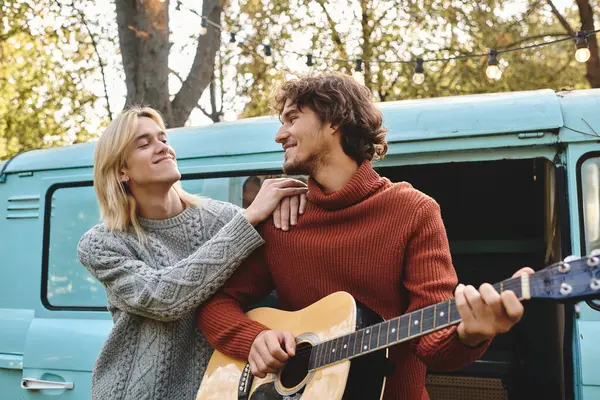 Two young and handsome people share a joyful moment as one plays guitar and the other gazes affectionately. Surrounded by nature, they radiate warmth and creativity during a sunny afternoon. — Stock Photo