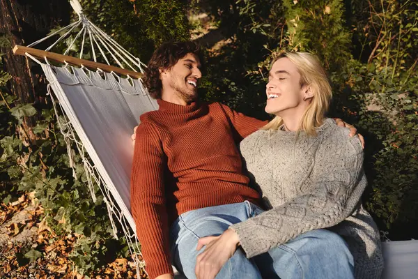 In a peaceful garden, two young men share laughter while relaxing on a hammock. Surrounded by lush greenery, their genuine smiles reflect a deep connection and happiness in each others presence. — Stock Photo