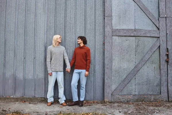 Under a serene afternoon sky, two young men stand side by side, holding hands and exchanging loving glances. Their cozy outfits and the weathered barn create an inviting atmosphere. — Stock Photo