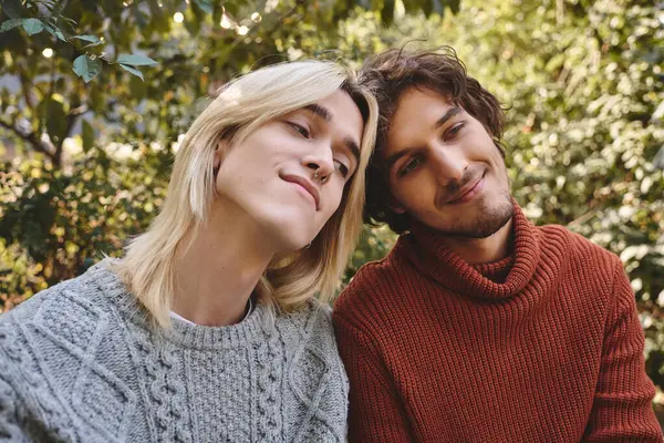 Young loving gay couple enjoy each others company in a lush garden filled with greenery. They wear cozy sweaters and share warm smiles, capturing a moment of friendship and affection under the sun. — Stock Photo