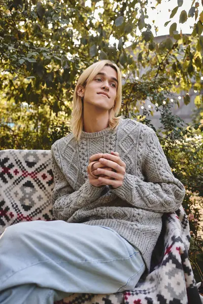 A young and handsome man relaxes on a patterned blanket outdoors, holding a warm drink with a thoughtful expression. Sunlight filters through green leaves, creating a peaceful atmosphere. — Stock Photo