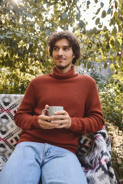 Relaxing in a lush outdoor setting, a young and handsome man holds a steaming cup while wearing a stylish sweater. Sunlight filters through the leaves, creating a serene atmosphere. — Stock Photo