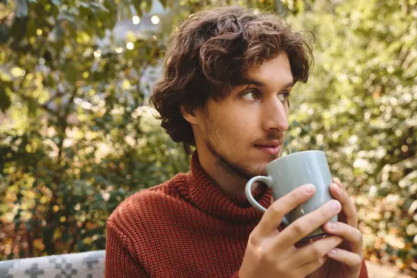 In a serene outdoor setting, a young and handsome man relaxes while holding a steaming coffee mug. Sunlight filters through the trees, creating a peaceful atmosphere. — Stock Photo