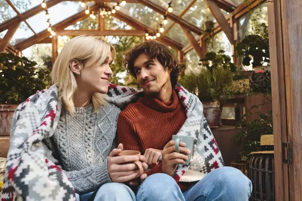 Due giovani e belli si siedono vicini, avvolti in un'accogliente coperta, godendo di un momento di pace in una serra splendidamente illuminata circondata da piante lussureggianti e verde. — Foto stock