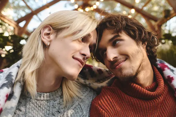 Dos jóvenes guapos comparten un momento tierno juntos, envueltos en una manta caliente en un hermoso espacio exterior adornado con luces suaves. Sus alegres expresiones reflejan un profundo vínculo y afecto. - foto de stock
