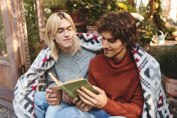 Two young men share an intimate moment wrapped in a cozy blanket. They are engrossed in a book, surrounded by lush greenery and a peaceful garden setting. A warm beverage adds to their comfort. — Stock Photo