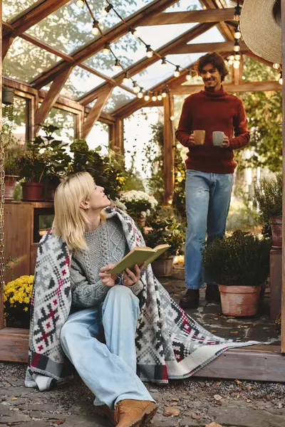 Young people enjoy a serene moment inside a greenhouse, surrounded by lush plants and flowers. One person reads a book while the other approaches with warm drinks, fostering a lively atmosphere. — Stock Photo