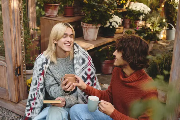 In a cozy garden adorned with plants and flowers, two young men enjoy each others company. One holds a cup while sharing a joyful story, wrapped in a blanket. — Stock Photo