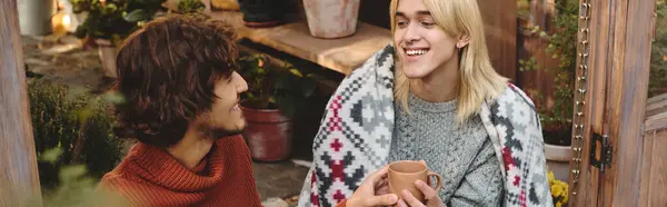 In a serene outdoor space adorned with plants, two young men share a warm conversation while wrapped in a stylish blanket. — Stock Photo