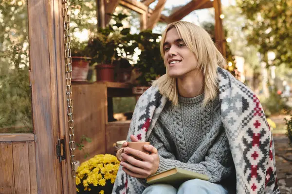 On a beautiful autumn day, a young, handsome person sits comfortably outside, wrapped in a warm blanket. — Stock Photo