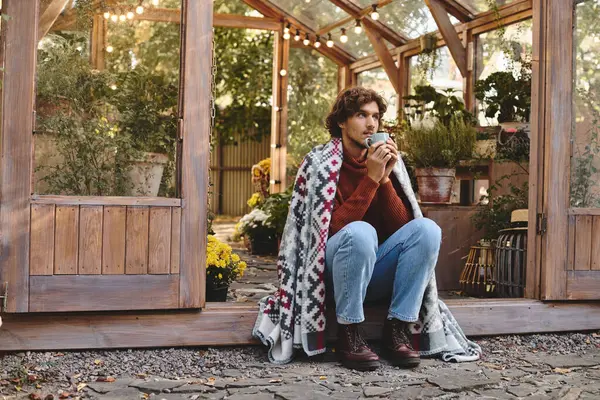 A young person relaxes outside a greenhouse, sipping a warm beverage. Surrounded by lush greenery and blooming flowers, they embody tranquility and style. — Stock Photo