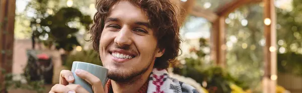 A young and handsome man sits contentedly in a lush garden, holding a warm cup and flashing a bright smile. Sunlight filters through greenery, enhancing the cheerful atmosphere. — Stock Photo