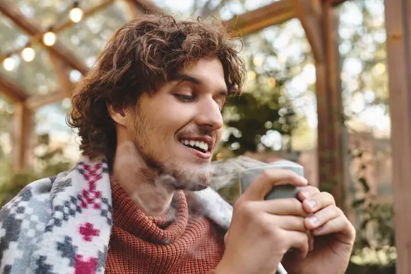 Enjoying a hot beverage while wrapped in a cozy blanket, a young man smiles brightly amidst a lush garden setting. Soft morning light filters through foliage, creating a tranquil atmosphere. — Stock Photo