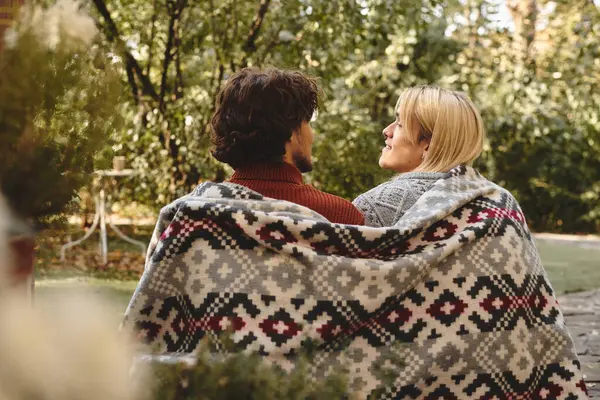 A young and handsome pair relax under a cozy blanket in a lush garden, enjoying each others company during a peaceful afternoon. — Stock Photo