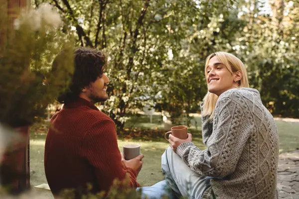 In a serene outdoor setting, Young loving gay couple with diverse styles relax together. With cups in hand and laughter in the air, they embody joy amidst lush greenery. — Stock Photo