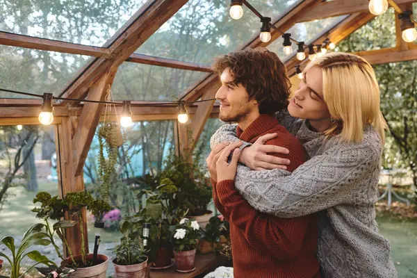 In a charming greenhouse illuminated by soft string lights, a young couple embraces intimately. Surrounded by lush greenery and potted plants, they exude warmth and connection, celebrating their love. — Stock Photo