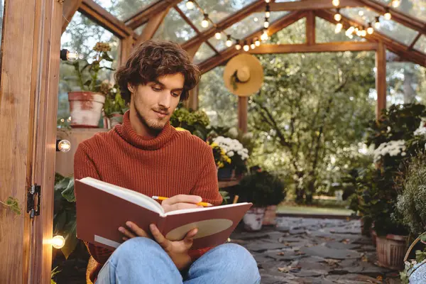 In a tranquil greenhouse filled with lush plants, a young man with stylish features is deeply engaged in writing. — Stock Photo