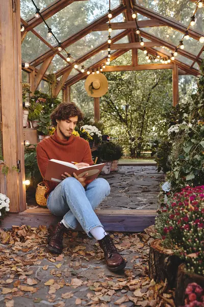 Seduto comodamente su un sentiero di pietra, un giovane uomo immerso in un libro gode dell'atmosfera tranquilla di una vibrante serra piena di fiori colorati e di luci calde.. — Foto stock