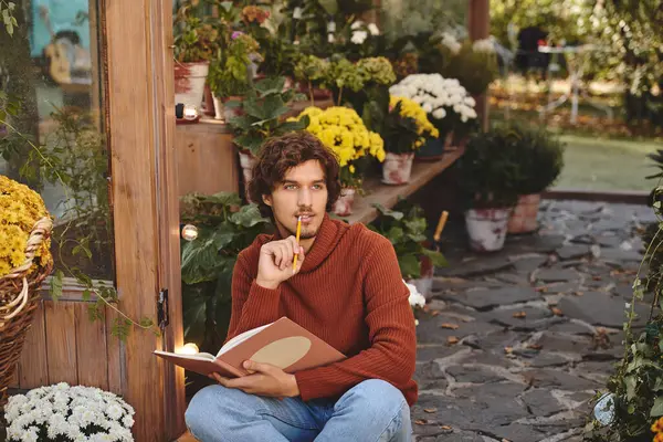 Surrounded by colorful flowers, a young and handsome man sits pensively with a notebook in hand. The setting is peaceful, inviting inspiration and reflection amidst natures beauty. — Stock Photo