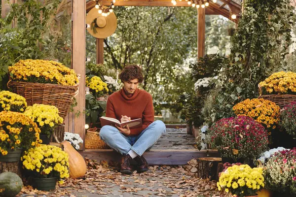 In a serene garden filled with blooming flowers, a young and handsome man sits on the ground, immersed in writing in a notebook. — Stock Photo