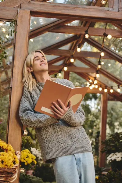 A young and attractive man relaxes in a tranquil greenhouse, surrounded by blooming flowers. Delighted by a book, he savors the cozy atmosphere amidst warm lights and greenery. — Stock Photo