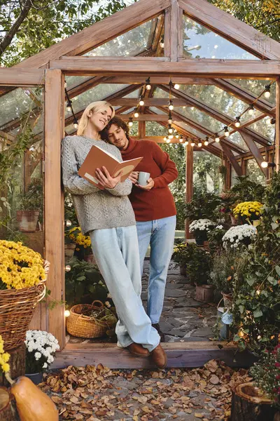En un invernadero bellamente decorado, un joven guapo se apoya sobre un marco de madera mientras su compañero lee un libro. El ambiente cálido se ve realzado por flores coloridas e iluminación suave. - foto de stock
