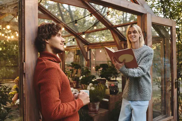 Two young and handsome friends share a delightful moment in a greenhouse. One reads joyfully while the other relaxes with a beverage. — Stock Photo