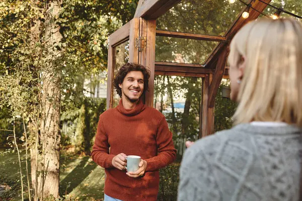 A young man wearing a cozy sweater smiles warmly while holding a cup in a vibrant garden. A cheerful exchange of ideas occurs amidst lush greenery and gentle sunlight. — Stock Photo