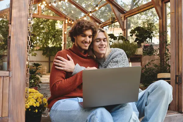 Two young men share a warm embrace as they sit together on the floor of a bright greenhouse. Surrounded by plants and flowers, they focus intently on their laptop, creating a peaceful atmosphere. — Stock Photo