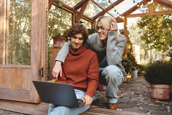 Two young men share a cozy moment at a garden space, laughing and looking at a laptop together. Surrounded by greenery, their joyful connection radiates warmth and happiness as they unwind. — Stock Photo
