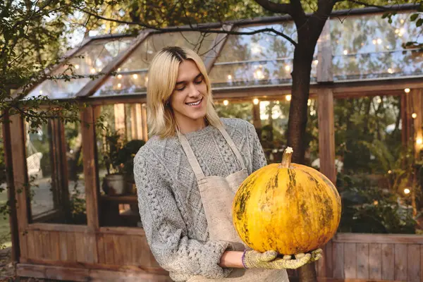 A charming young man with long blond hair holds a vibrant pumpkin with a warm smile. — Stock Photo