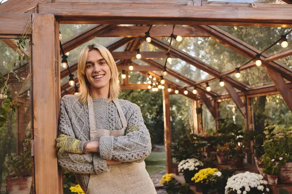 A young man stands in a cozy greenhouse adorned with warm lights, surrounded by lush plants. Their smile radiates happiness while they showcase their love for gardening. — Stock Photo