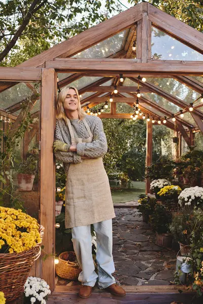 In a serene garden filled with colorful blooms, a young man in a cozy sweater and apron leans against a wooden frame, appreciating the beauty around him. — Stock Photo