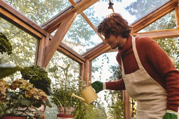In a peaceful greenhouse filled with lush greenery, a young and handsome man lovingly waters potted plants. Sunlight filters through the glass, creating a tranquil atmosphere. — Stock Photo