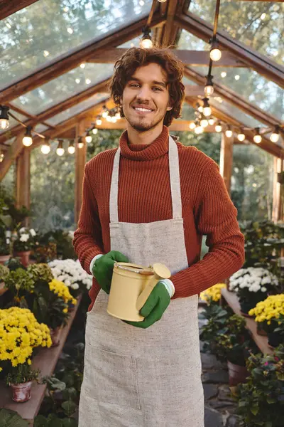 In a warm and inviting greenhouse, a young man with a joyful expression holds a yellow pot while dressed in a cozy sweater and apron. Lush flowers surround him, capturing the beauty of nature. — Stock Photo