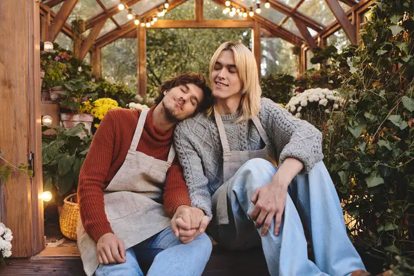 Two young men sit closely together in a charming greenhouse, surrounded by vibrant flowers. They share a tender moment, showcasing love and connection, while warm lights illuminate the lush greenery. — Stock Photo