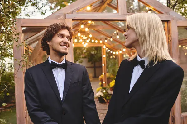 Two young men in formal attire share a joyful moment in a beautifully lit outdoor setting. The enchanting garden is adorned with twinkling lights, creating a romantic atmosphere for their celebration. — Stock Photo