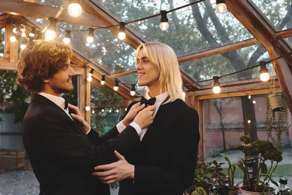 In a charming greenhouse adorned with fairy lights, a young couple enjoys a playful moment as one adjusts the others bow tie. — Stock Photo