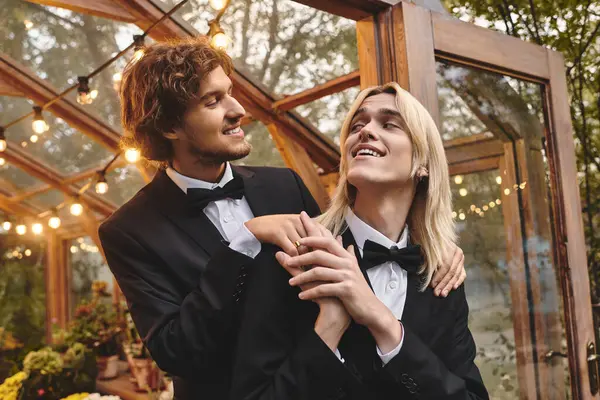 In a beautifully lit garden, two young men dressed in elegant tuxedos share a joyful moment. Surrounded by nature and soft lights, their laughter and connection shine brightly in the evening ambiance. — Stock Photo
