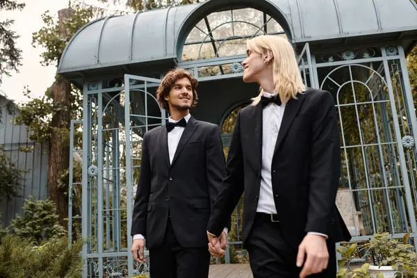 In a charming garden adorned with greenery, two young men dressed in elegant suits walk hand in hand. Their cheerful expressions capture the essence of love and celebration in a romantic atmosphere. — Stock Photo