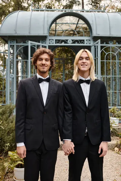 In un bel giardino, due giovani in abiti eleganti si tengono per mano e sorridono, irradiando felicità. Circondato da un verde lussureggiante e una struttura affascinante, il loro collegamento è commovente.. — Foto stock
