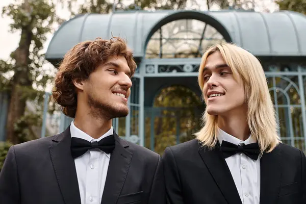 Two young men dressed in stylish black suits share a joyful connection in a beautifully landscaped garden. — Stock Photo