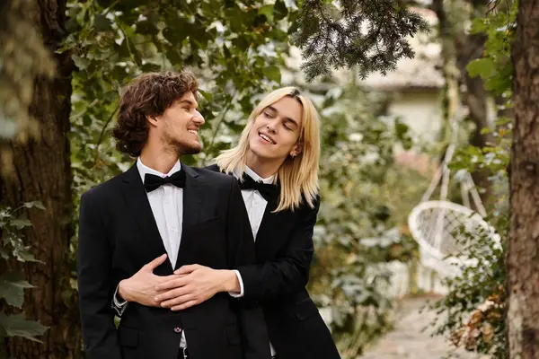 Two handsome young men embrace playfully, dressed in stylish black suits with bow ties, surrounded by lush greenery. — Stock Photo