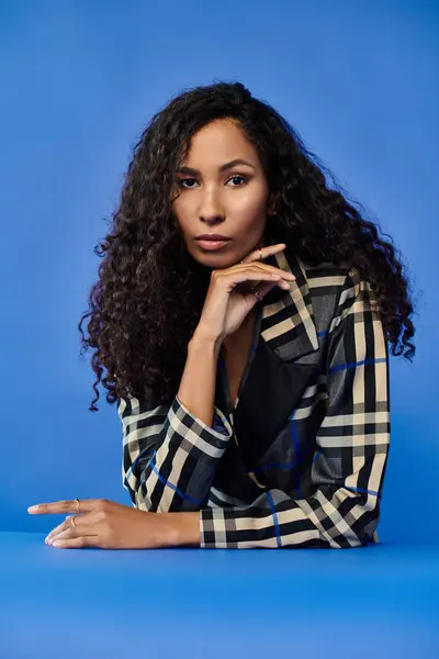 Una donna sicura di sé con i capelli ricci posa pensieroso su uno sfondo blu. — Foto stock