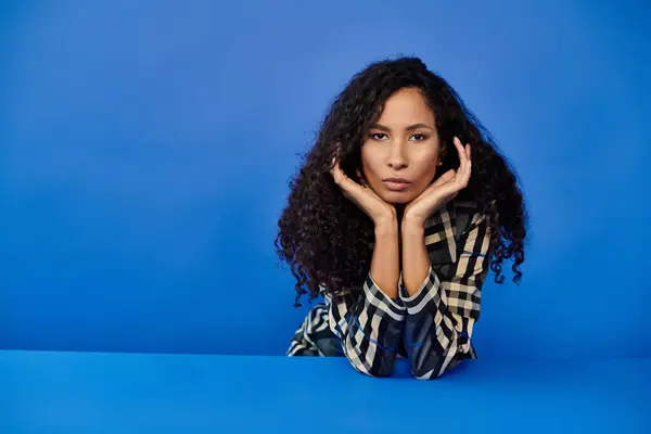 Captivating woman gazes thoughtfully while resting her chin on her hands, set against blue. — Stock Photo