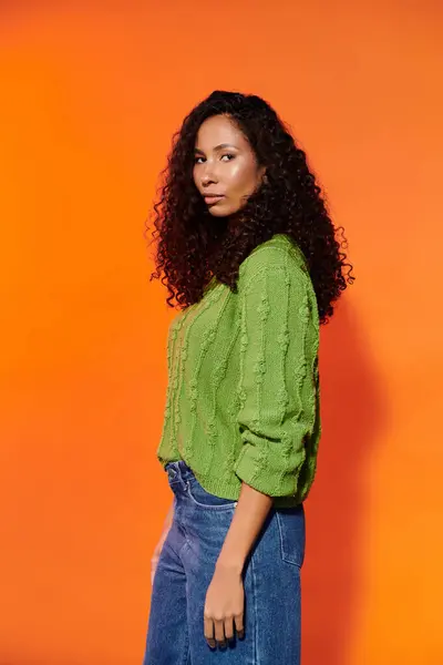 A woman with beautiful curly hair poses confidently in a stylish green sweater against orange. — Stock Photo