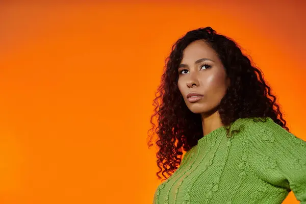 A confident woman in a green sweater poses gracefully against an orange backdrop. — Stock Photo