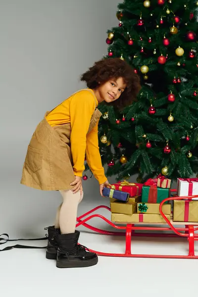 Il piccolo sorride mentre prepara una slitta colorata vicino a un albero splendidamente decorato. — Foto stock