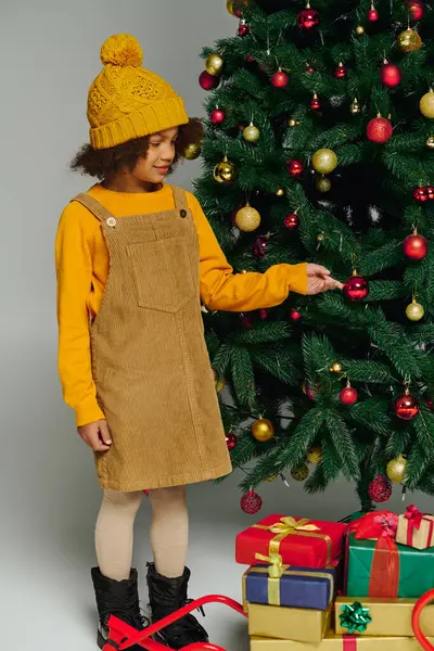A joyful girl adds a shiny ornament to a vibrant Christmas tree surrounded by gifts. — Stock Photo